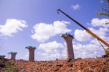 A construction site featuring several tall concrete pillars, with a crane positioned on the right side lifting materials. The sky is bright blue with scattered fluffy white clouds. The ground is rough with dirt and rocks, and green mesh is visible on top of the pillars.