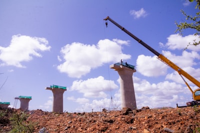 A construction site featuring several tall concrete pillars, with a crane positioned on the right side lifting materials. The sky is bright blue with scattered fluffy white clouds. The ground is rough with dirt and rocks, and green mesh is visible on top of the pillars.