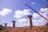 A construction site featuring several tall concrete pillars, with a crane positioned on the right side lifting materials. The sky is bright blue with scattered fluffy white clouds. The ground is rough with dirt and rocks, and green mesh is visible on top of the pillars.