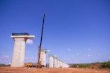 Tall concrete bridge pillars under construction stand in a line on a clear day. A large yellow crane is positioned beside one of the pillars, and a few workers can be seen at the top of the pillars. The background shows a clear blue sky with some clouds, and the ground is a dry, earthy surface.