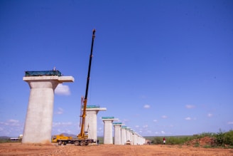 Tall concrete bridge pillars under construction stand in a line on a clear day. A large yellow crane is positioned beside one of the pillars, and a few workers can be seen at the top of the pillars. The background shows a clear blue sky with some clouds, and the ground is a dry, earthy surface.