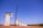 Tall concrete bridge pillars under construction stand in a line on a clear day. A large yellow crane is positioned beside one of the pillars, and a few workers can be seen at the top of the pillars. The background shows a clear blue sky with some clouds, and the ground is a dry, earthy surface.