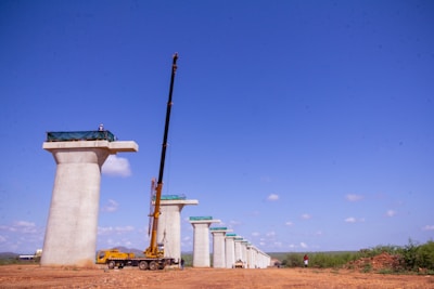 Tall concrete bridge pillars under construction stand in a line on a clear day. A large yellow crane is positioned beside one of the pillars, and a few workers can be seen at the top of the pillars. The background shows a clear blue sky with some clouds, and the ground is a dry, earthy surface.