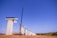 Tall concrete bridge pillars under construction stand in a line on a clear day. A large yellow crane is positioned beside one of the pillars, and a few workers can be seen at the top of the pillars. The background shows a clear blue sky with some clouds, and the ground is a dry, earthy surface.