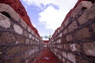 A stone trench with sturdy walls stretches into the distance. The sky above is partially cloudy, casting distinct shadows and light patterns on the stones. The deep red earth on the sides contrasts sharply with the grey stones. In the distance, a person in work attire stands atop the trench, holding a long pole.