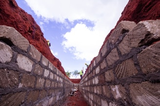 A deep trench lined with large, rough stone blocks, with a vivid red earthen wall on either side. The view looks upward towards a blue sky with scattered clouds. Workers in safety gear, including helmets and reflective vests, are visible near the top of the trench.