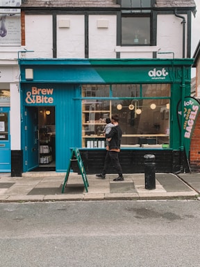 A quaint storefront with blue and teal accents features signs for 'Brew & Bite' and 'dot. real good bagels.' A person in dark clothing, holding a child, walks past on the sidewalk. Inside, warm lighting and shelves with products create a cozy atmosphere. A small signboard is placed on the pavement near the entrance.