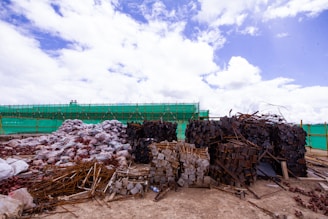 Large piles of construction materials and debris are scattered across a barren site under a partly cloudy sky. The materials include metal rods, stacked wooden planks, and heaps of plastic-wrapped bundles. A green mesh fence runs along the background, indicating construction or renovation work.