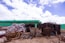 Large piles of construction materials and debris are scattered across a barren site under a partly cloudy sky. The materials include metal rods, stacked wooden planks, and heaps of plastic-wrapped bundles. A green mesh fence runs along the background, indicating construction or renovation work.