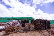 Large piles of construction materials and debris are scattered across a barren site under a partly cloudy sky. The materials include metal rods, stacked wooden planks, and heaps of plastic-wrapped bundles. A green mesh fence runs along the background, indicating construction or renovation work.