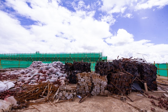 Large piles of construction materials and debris are scattered across a barren site under a partly cloudy sky. The materials include metal rods, stacked wooden planks, and heaps of plastic-wrapped bundles. A green mesh fence runs along the background, indicating construction or renovation work.