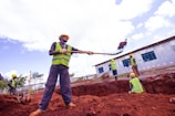 Construction workers are actively engaged in digging and moving soil at a construction site. They are wearing safety gear, including helmets and reflective vests, and using shovels. One worker is in the foreground lifting a load with a shovel, while others work in the background near a corrugated metal building. The site appears to be outdoors with a clear sky.