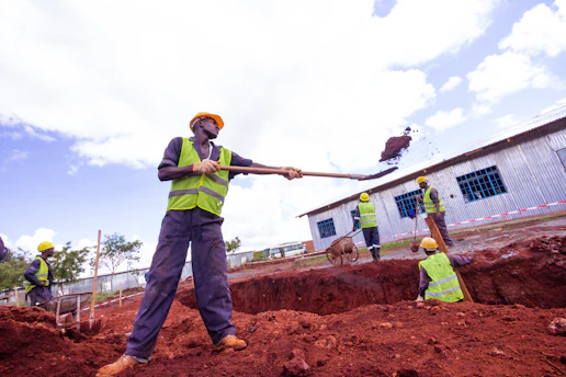 Construction workers are actively engaged in digging and moving soil at a construction site. They are wearing safety gear, including helmets and reflective vests, and using shovels. One worker is in the foreground lifting a load with a shovel, while others work in the background near a corrugated metal building. The site appears to be outdoors with a clear sky.