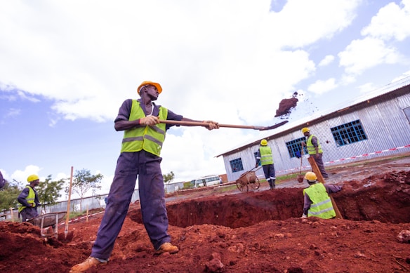 Construction workers are actively engaged in digging and moving soil at a construction site. They are wearing safety gear, including helmets and reflective vests, and using shovels. One worker is in the foreground lifting a load with a shovel, while others work in the background near a corrugated metal building. The site appears to be outdoors with a clear sky.