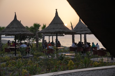 A seaside setting at sunset with people gathered under thatched umbrellas. The scene includes lush greenery and flowering plants, creating a tropical ambiance. The sun is partially visible near the horizon over the water, adding a warm glow to the scene.