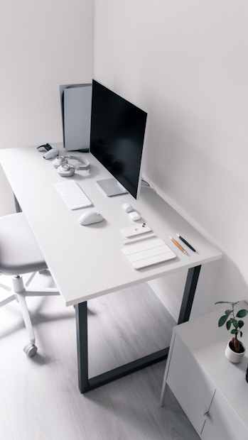 A minimalist and clean workspace features a sleek white desk with a computer monitor, keyboard, mouse, and a set of wireless headphones. A gaming console is placed to the left, and several neatly arranged stationery items such as notebooks and pens are present. A potted plant is situated in the corner on a small cabinet.