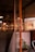 A cozy interior with warm lighting features wooden and concrete elements. A potted plant is placed near a glass partition, and various shelves are visible in the background along with hanging lights and fans. The ambiance suggests a relaxed environment, possibly a cafe or lounge area.