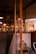 A cozy interior with warm lighting features wooden and concrete elements. A potted plant is placed near a glass partition, and various shelves are visible in the background along with hanging lights and fans. The ambiance suggests a relaxed environment, possibly a cafe or lounge area.
