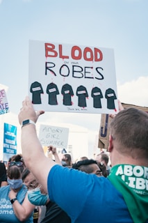 A group of people gathered in a protest. One prominent sign reads 'BLOOD ON YOUR ROBES' with illustrations of black robes dripping red. Another smaller sign says 'ANGRY GIRL SUMMER.' The crowd includes individuals wearing masks and holding various signs. The sky is clear blue in the background.