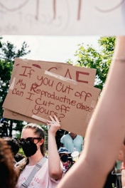 A person wearing sunglasses and a face mask holds a cardboard sign with a message about reproductive rights, surrounded by other individuals. The scene takes place outdoors, with green foliage visible in the background and sunlight casting shadows.