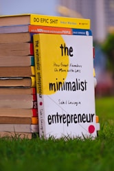A stack of books is placed on green grass. The book in the foreground is titled 'The Minimalist Entrepreneur' by Sahil Lavingia, with a yellow and white cover. Other books in the stack include 'Do Epic Shit' by Ankur Warikoo and 'How to Win Friends and Influence People' by Dale Carnegie. The background is blurred, indicating an outdoor setting.