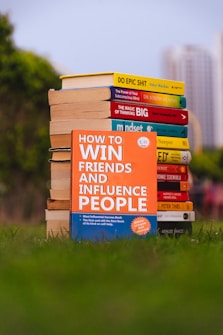 A stack of self-help books arranged outdoors, with vibrant covers. The central focus is on 'How to Win Friends and Influence People' prominently displayed in the foreground. The books are placed on lush green grass, with blurred city buildings in the background.