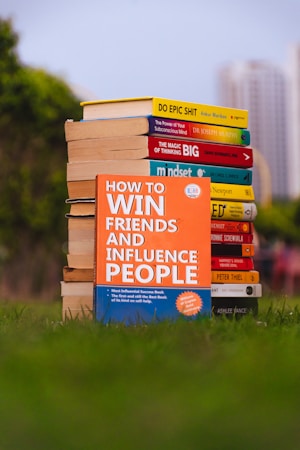 A stack of self-help books arranged outdoors, with vibrant covers. The central focus is on 'How to Win Friends and Influence People' prominently displayed in the foreground. The books are placed on lush green grass, with blurred city buildings in the background.