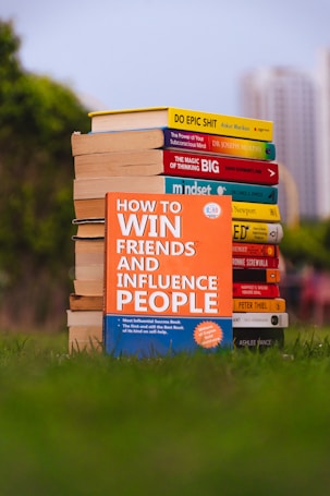 A stack of self-help books arranged outdoors, with vibrant covers. The central focus is on 'How to Win Friends and Influence People' prominently displayed in the foreground. The books are placed on lush green grass, with blurred city buildings in the background.