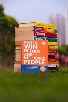 A stack of self-help books arranged outdoors, with vibrant covers. The central focus is on 'How to Win Friends and Influence People' prominently displayed in the foreground. The books are placed on lush green grass, with blurred city buildings in the background.