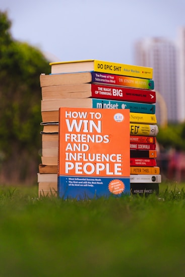 A stack of self-help books arranged outdoors, with vibrant covers. The central focus is on 'How to Win Friends and Influence People' prominently displayed in the foreground. The books are placed on lush green grass, with blurred city buildings in the background.