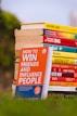 A stack of colorful self-help and motivational books is placed on grass, with titles clearly visible. The books are arranged with one book prominently displayed in the foreground, titled 'How to Win Friends and Influence People'. The setting appears to be outdoors with a blurred backdrop of greenery and a clear sky.