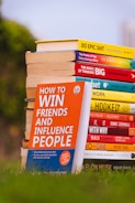 A stack of colorful self-help and motivational books is placed on grass, with titles clearly visible. The books are arranged with one book prominently displayed in the foreground, titled 'How to Win Friends and Influence People'. The setting appears to be outdoors with a blurred backdrop of greenery and a clear sky.