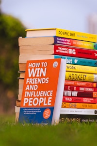 A stack of colorful self-help and motivational books is placed on grass, with titles clearly visible. The books are arranged with one book prominently displayed in the foreground, titled 'How to Win Friends and Influence People'. The setting appears to be outdoors with a blurred backdrop of greenery and a clear sky.