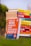 A stack of colorful self-help and motivational books is placed on grass, with titles clearly visible. The books are arranged with one book prominently displayed in the foreground, titled 'How to Win Friends and Influence People'. The setting appears to be outdoors with a blurred backdrop of greenery and a clear sky.