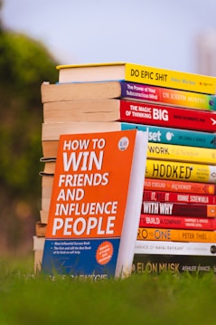 A stack of colorful self-help and motivational books is placed on grass, with titles clearly visible. The books are arranged with one book prominently displayed in the foreground, titled 'How to Win Friends and Influence People'. The setting appears to be outdoors with a blurred backdrop of greenery and a clear sky.
