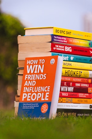 A stack of colorful self-help and motivational books is placed on grass, with titles clearly visible. The books are arranged with one book prominently displayed in the foreground, titled 'How to Win Friends and Influence People'. The setting appears to be outdoors with a blurred backdrop of greenery and a clear sky.
