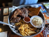 A plate of barbecue ribs with a side of crispy French fries, a small cup of dipping sauce, a dish of creamy coleslaw, and a piece of toasted bread. The food is served on a round tray with a checkered paper liner.