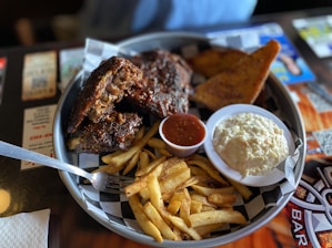 A plate of barbecue ribs with a side of crispy French fries, a small cup of dipping sauce, a dish of creamy coleslaw, and a piece of toasted bread. The food is served on a round tray with a checkered paper liner.
