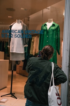 A person dressed in a dark green jacket stands outside a storefront window, apparently taking a photo or inspecting the display. The display features two mannequins; one is dressed in a white t-shirt with a gray cardigan and the other in a vibrant green shirt. Bold blue lettering on the window reads 'AMERICAN VINTAGE S/S 22 SOLDES'. The person carries a white tote bag with red text.
