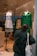 A person dressed in a dark green jacket stands outside a storefront window, apparently taking a photo or inspecting the display. The display features two mannequins; one is dressed in a white t-shirt with a gray cardigan and the other in a vibrant green shirt. Bold blue lettering on the window reads 'AMERICAN VINTAGE S/S 22 SOLDES'. The person carries a white tote bag with red text.