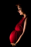 A pregnant woman stands gracefully, illuminated by soft lighting against a dark background. She wears a vibrant red dress that accentuates her belly, and a floral headpiece rests on her head. Her gaze is gentle and focused downward.