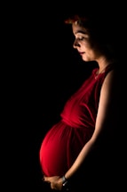A pregnant woman stands gracefully, illuminated by soft lighting against a dark background. She wears a vibrant red dress that accentuates her belly, and a floral headpiece rests on her head. Her gaze is gentle and focused downward.