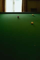 A green billiard table with three balls positioned on it, including a red ball in the center and two yellow balls at different angles. There is a window in the background that casts natural light onto the scene.