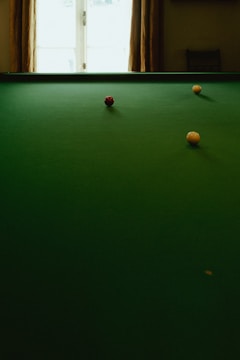 A green billiard table with three balls positioned on it, including a red ball in the center and two yellow balls at different angles. There is a window in the background that casts natural light onto the scene.