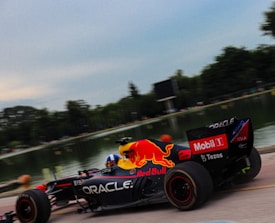 A Formula 1 race car with Red Bull branding speeds along a racetrack beside a body of water. The background features blurred greenery, suggesting high speed.