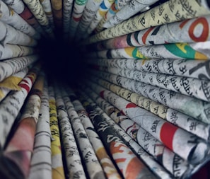 A perspective view of rolled newspapers creating a tunnel-like effect. The papers contain various texts and colorful images, featuring languages that appear to be in Hindi. The vibrant assortment of texts and colors form a visually engaging pattern as the rolls converge towards a dark center.