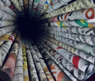 A perspective view of rolled newspapers creating a tunnel-like effect. The papers contain various texts and colorful images, featuring languages that appear to be in Hindi. The vibrant assortment of texts and colors form a visually engaging pattern as the rolls converge towards a dark center.