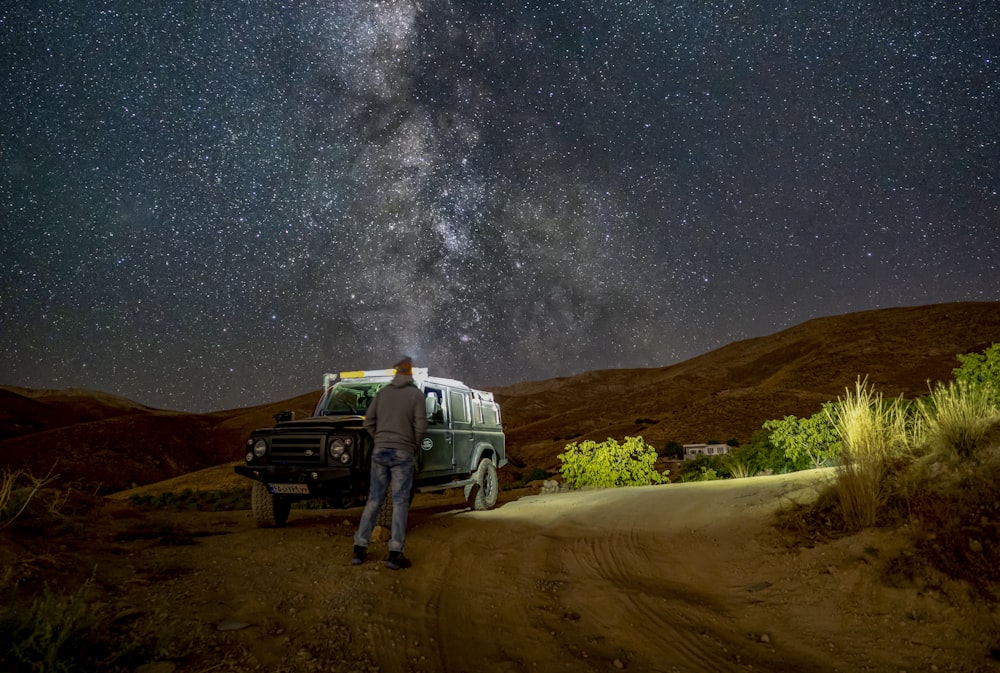 a person standing next to a car on a dirt road with stars in the sky