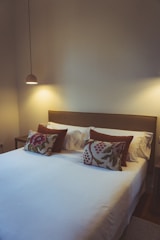 A cozy, minimalist bedroom with a neatly made king-sized bed. The bed features white linens and is adorned with two patterned pillows with floral designs. Two soft, brown accent pillows rest behind them. A simple, modern pendant light hangs from the ceiling, casting a warm glow over the bedside area. Neutral walls contribute to the calming atmosphere.