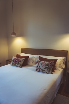 A cozy, minimalist bedroom with a neatly made king-sized bed. The bed features white linens and is adorned with two patterned pillows with floral designs. Two soft, brown accent pillows rest behind them. A simple, modern pendant light hangs from the ceiling, casting a warm glow over the bedside area. Neutral walls contribute to the calming atmosphere.
